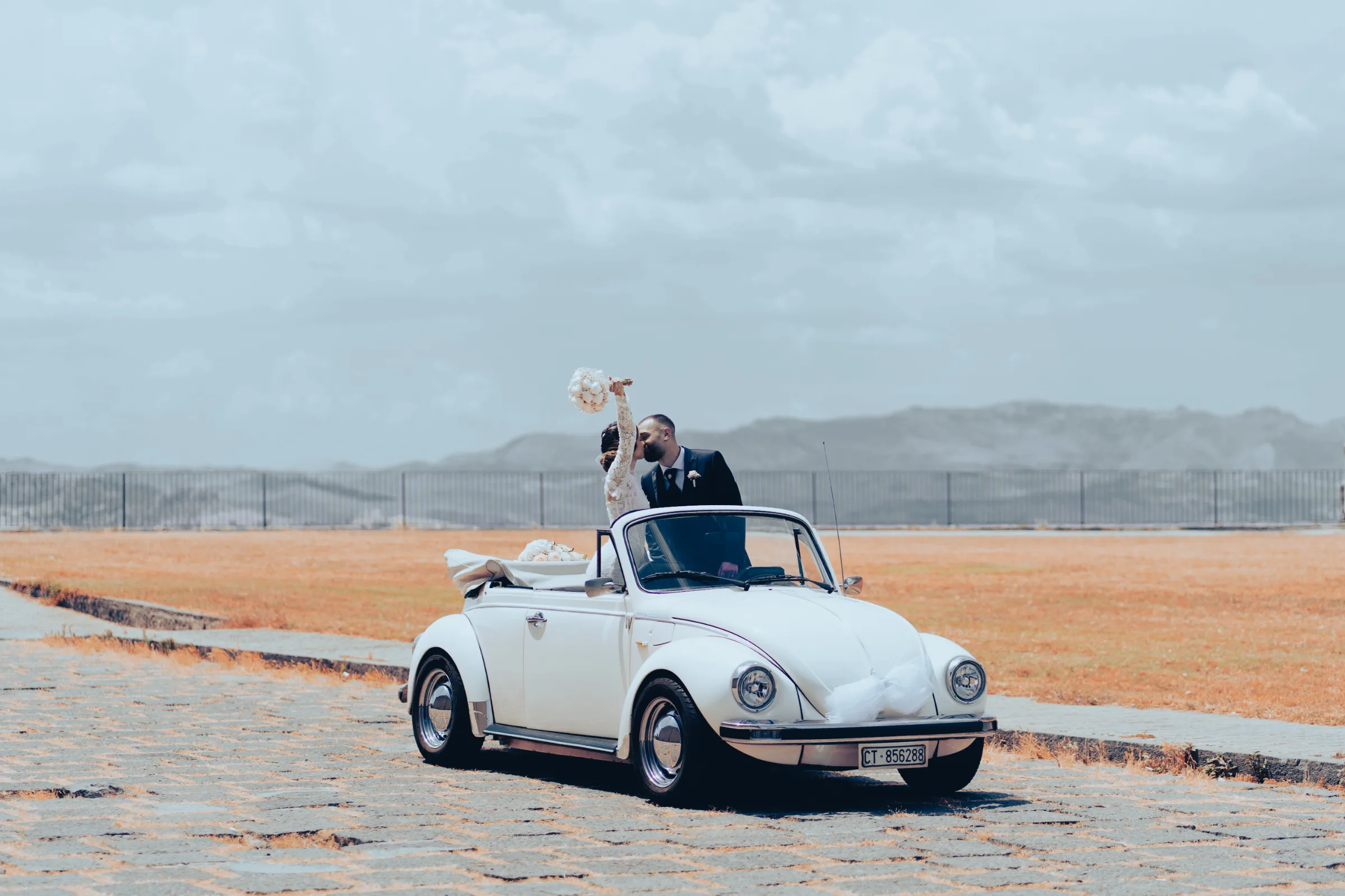 Newlywed couple standing up and sharing a kiss in a classic convertible Beetle overlooking a picturesque Sicilian countryside valley